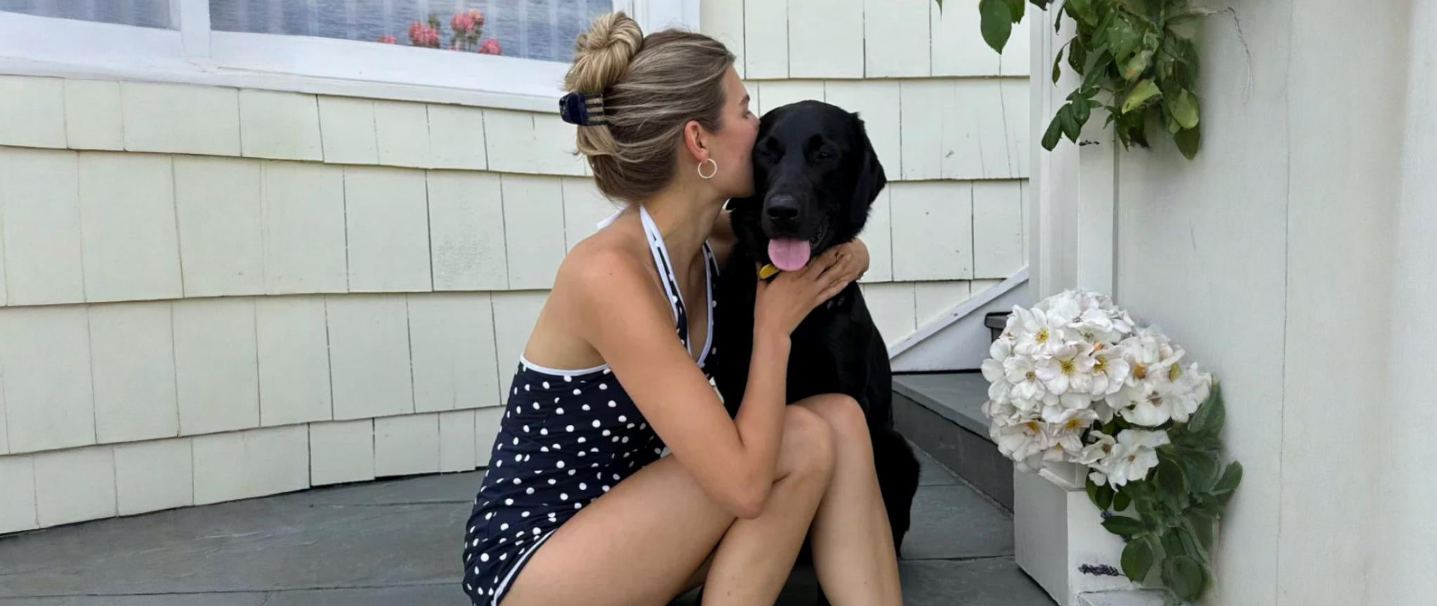 Woman sitting on steps with a black dog, surrounded by flowers and plants.